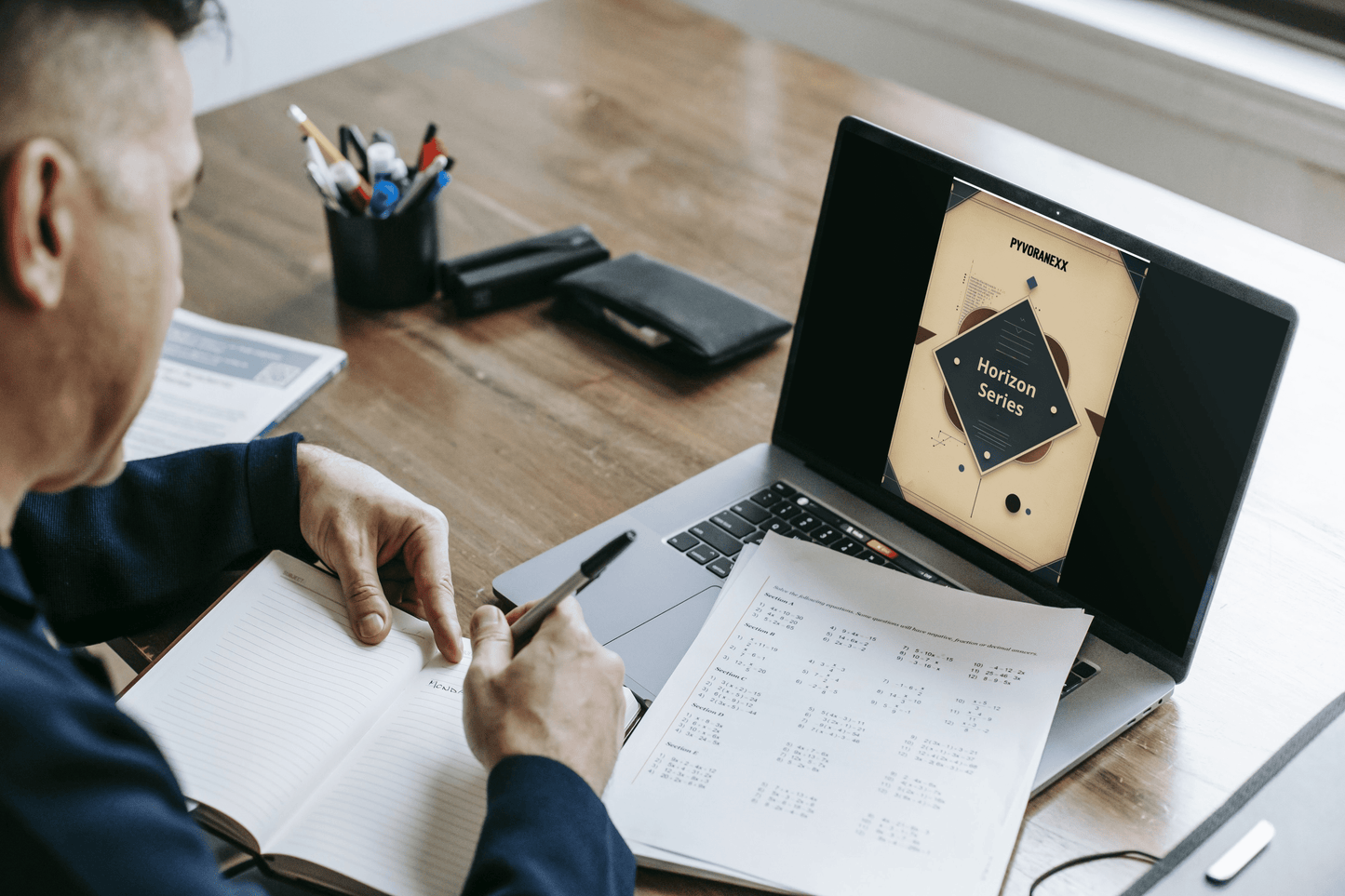 Person working at a desk with a laptop and notebook, surrounded by office supplies.