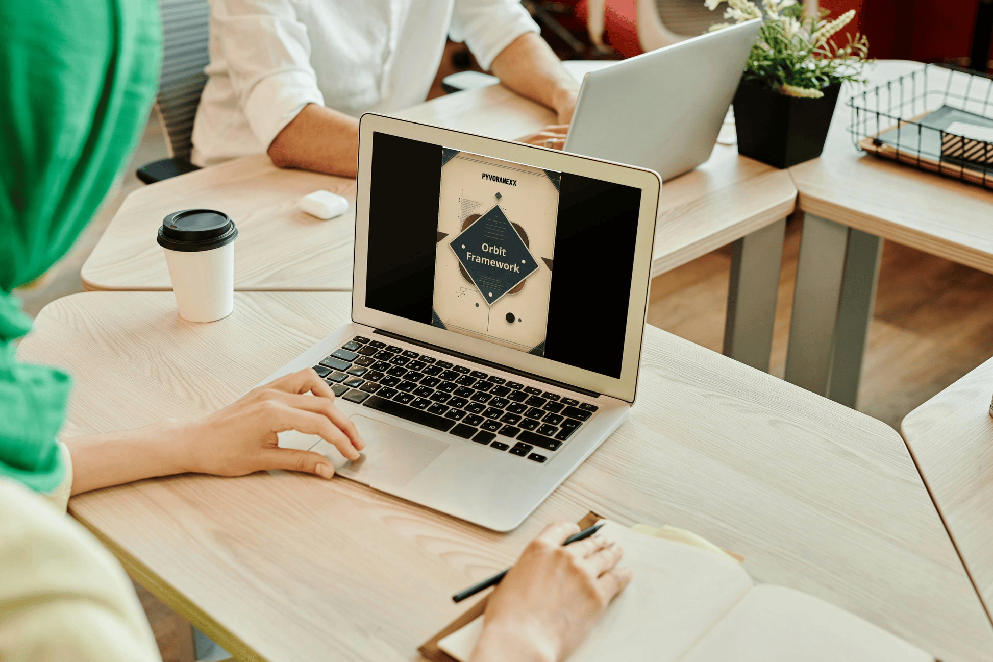 Person using a laptop at a desk with another person in the background.