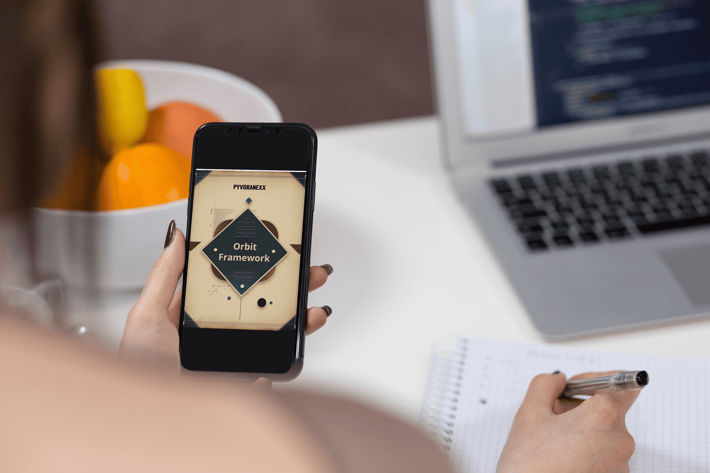 Person holding a phone with Python course on a desk with a laptop and fruit bowl.