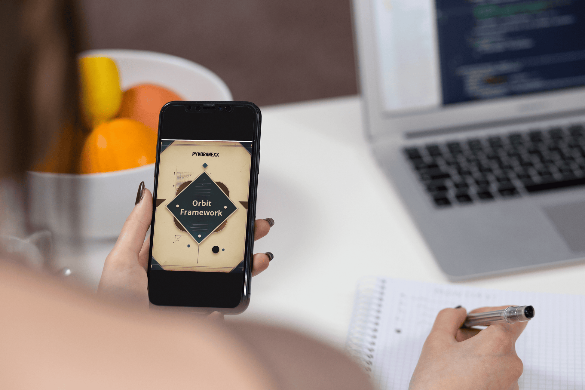 Person holding a phone with Python course on a desk with a laptop and fruit bowl.